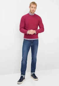 Bordeauxroter Pullover über einem blauen, geblümten Hemd, kombiniert mit blauen Jeans und marineblauen Turnschuhen. Der Pullover hat einen Rundhalsausschnitt und gerippte Bündchen.