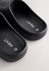 Schwarze Slides mit strukturierter Oberfläche, abgerundeter Form und breiten Riemen. Auf der Innensohle sind das Logo "NEXXT" und die Größe "12" zu sehen.