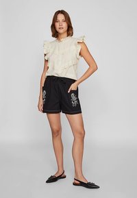 Světlá béžová bezrukávová halenka s volánovými patry a krajkovou lemovkou, kombinovaná s černými šortkami s květinovou výšivkou a stahováním v pase.