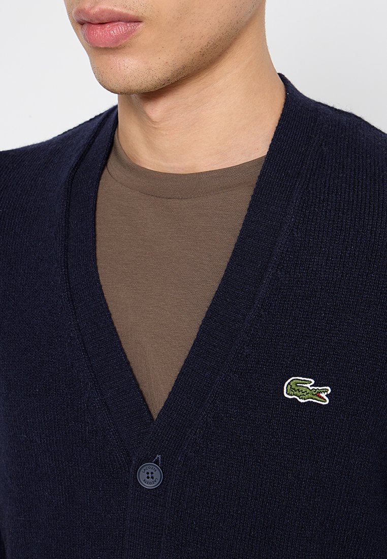 Cardigan blu navy con lavorazione a maglia testurizzata, design a V e chiusura con un bottone. Presenta un piccolo logo di coccodrillo verde sul petto.
