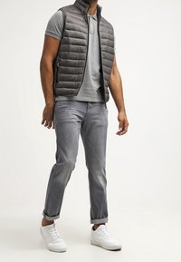 Tummanharmaa toppaliivi, lyhyet hihat ja korkea kaulus. Alla harmaa poolopaita, haalistuneet harmaat farkut ja valkoiset tennarit.