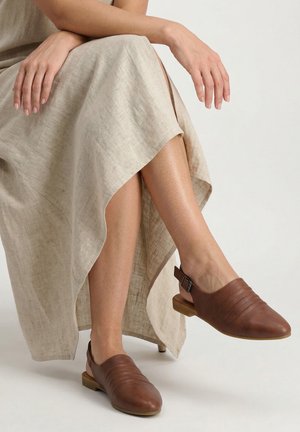 Vrouw zittend, draag een beige linnen rok en bruine leren slingback platte schoenen met gesp op een neutrale achtergrond.