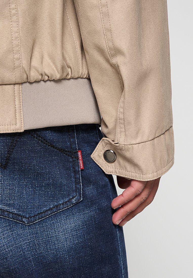 Veste beige avec taille élastique et poignets, texture semblable à du daim, dotée d'un bouton-pression. Portée sur un jean en denim foncé avec une étiquette rouge.