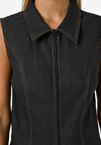 Svart ärmlös topp med dragkedja fram, med kontrasterande orange sömmar och klassisk krage. Slät textur, figursydd design.