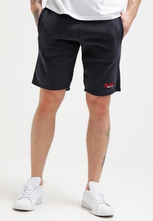 Mężczyzna ubrany w czarne krótkie spodenki do kolan, białe buty sportowe i białą koszulę, stoi z rękami w kieszeniach na jednolitym tle.