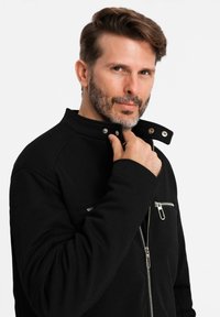 Veste zippée noire avec col montant, dotée d'une poche avant et de détails en métal. Le tissu texturé apporte de la profondeur au design.