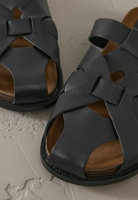 Zwarte leren sandalen met kruislings gespen, open teen ontwerp en een bruine, gestructureerde binnenzool. Stevige zwarte rubberen zool voor grip.