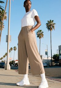 Fiatal személy áll napos utcán, magas pálmafák között, fehér pólót, bézs színű, bő szárú nadrágot, fehér zoknit és fehér tornacipőt visel.