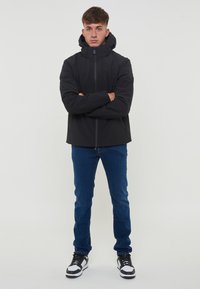 Giacca nera impermeabile con colletto alto e cappuccio regolabile; presenta una zip frontale e due tasche laterali. Indossata con jeans in denim blu.