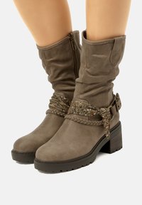 Botas de ante marrón hasta la rodilla con una correa trenzada y una hebilla decorativa, que presentan un acabado texturizado y un tacón grueso.