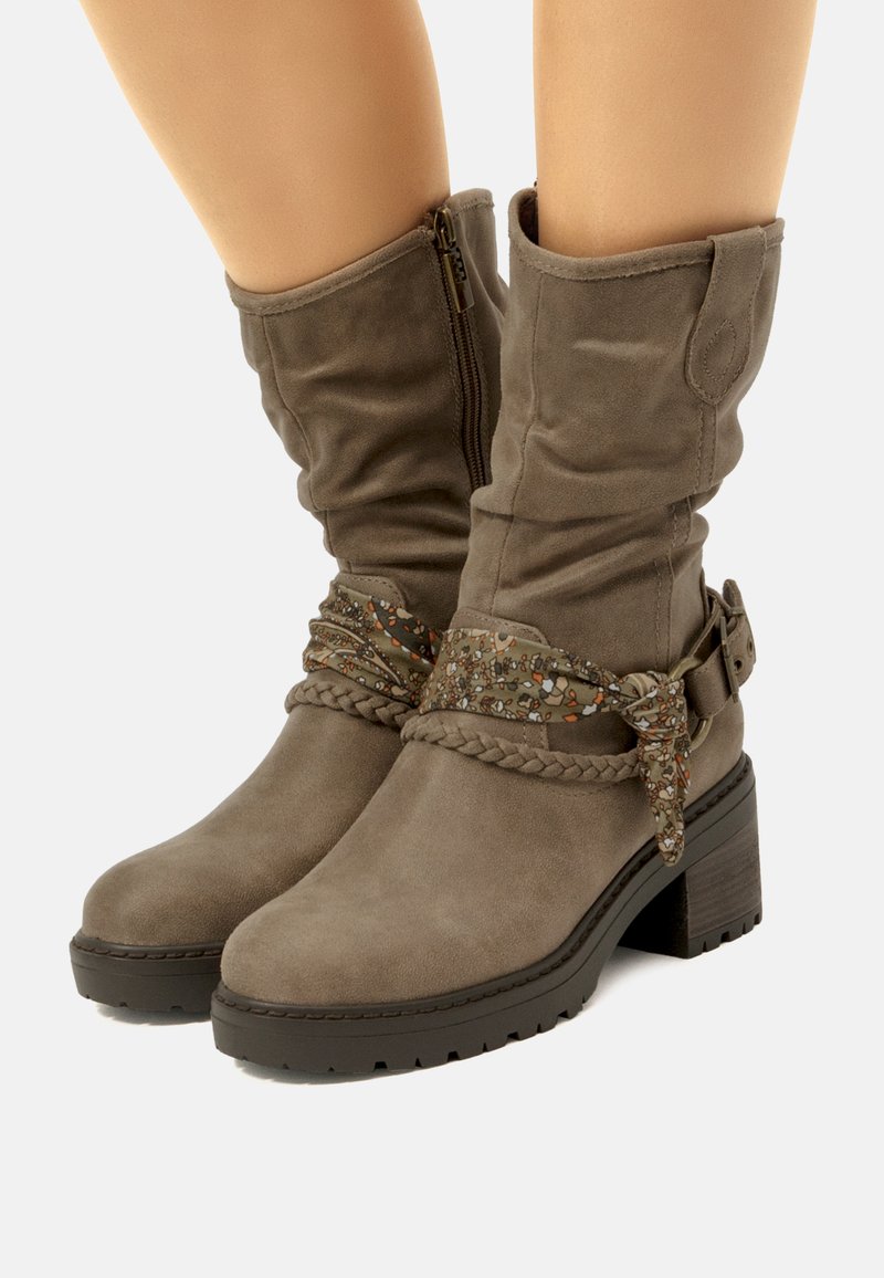 Botas de ante marrón hasta la rodilla con una correa trenzada y una hebilla decorativa, que presentan un acabado texturizado y un tacón grueso.