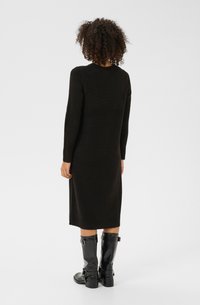 Robe en tricot noire avec manches longues, poignets côtelés et ourlet mi-long. Associée à des bottes noires brillantes jusqu'aux genoux avec des boucles.