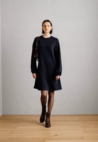 Donker marineblauwe jurk met lange mouwen, een aangepaste lijfje en een uitlopende zoom, gecombineerd met bronzen enkellaarsjes. Gladde textuur en minimalistisch ontwerp.