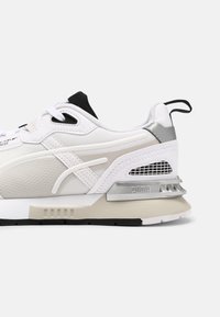 Vue latérale d'une basket Puma blanche et gris clair avec des accents noirs, une semelle texturée et un onglet à l'arrière.
