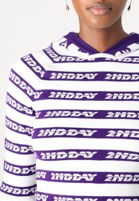 Pullover con cappuccio a righe bianche e viola. Presenta il testo ripetuto "2NDDAY" in grassetto. Materiale morbido a maglia con scollatura a coste.