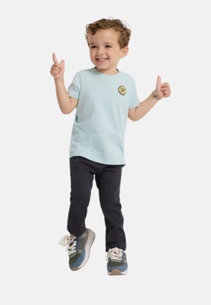 Camiseta de algodón azul claro con un logo amarillo circular, combinada con pantalones oscuros y zapatillas multicolor. De pie con los pulgares en alto.