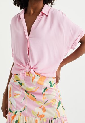 Kvinde iført en lyserød skjorte med knapper, bundet ved taljen, og et blomsterprintet nederdel med orange, gule, grønne og pink mønstre.