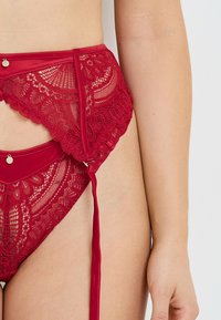 Lingerie in pizzo rosso con motivi floreali, dettagli in raso, fiocchi laterali e orlo smerlato. Dotata di parti in metallo per la chiusura.