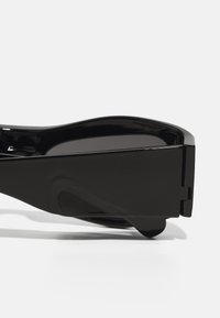 Lunettes de soleil noires avec une finition brillante, design de monture angulaire, verres foncés et une branche sculptée distinctive pour une meilleure prise en main.