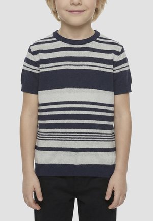 Enfant portant un pull rayé bleu marine et gris à manches courtes et un pantalon noir, debout devant un fond clair uni.