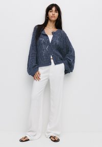 Cardigan en tricot bleu marine avec motif géométrique, fermeture à boutons et manches volumineuses ; associé à un pantalon large blanc et des tongs noires.