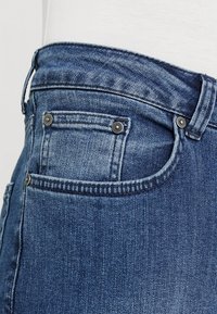 Jeans i denim med medelhög midja, blåtvätt, standardfickor och kontrasterande sömmar. Har metalldetaljer på bakfickorna.