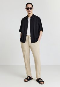 Uomo che indossa una camicia nera a maniche corte con bottoni, una maglietta bianca, pantaloni beige sgualciti, sandali neri e occhiali da sole, in piedi con le mani in tasca.