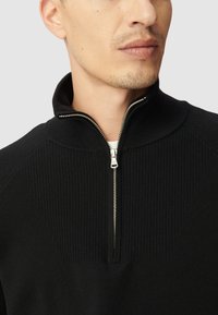 Czarny sweter dzianinowy z zamkiem błyskawicznym i stójką, o ribbowanej teksturze na ramionach i gładkiej części ciała. Zauważono srebrny detal zamka.