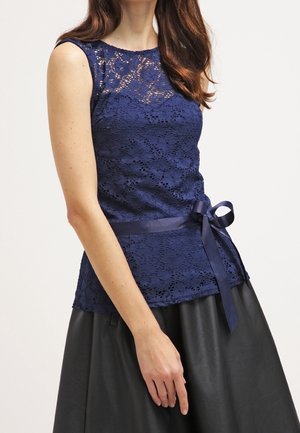 Marineblauw kanttop met een getailleerde lijfje, bloemenpatroon, korte mouwen en een satijnen strik bij de taille. Gecombineerd met een zwarte rok.