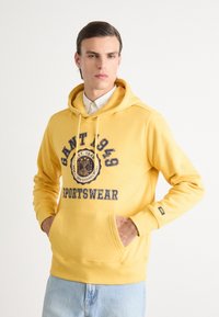 Hoodie jaune avec une poche frontale, présentant un motif graphique circulaire et un design textuel en bleu marine. Matière douce avec une coupe régulière et des cordons de serrage ajustables.