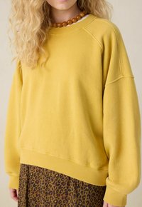 Femme portant un sweat-shirt jaune ample, un collier en perles marron et une jupe à imprimé léopard marron sur un fond neutre.