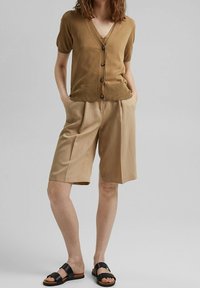Brun kortärmad cardigan med knappar, matchad med beige veckade shorts. Svarta sandaler med remmar fulländar looken. Enkelt, avslappnat design.