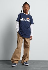 Dunkelblaues T-Shirt mit weißem "ellesse"-Logo, das über einem weißen Langarmshirt getragen wird; kombiniert mit beigen Cargo-Hosen und dunkelblauen Sneakers.