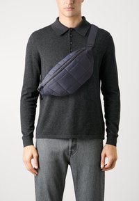 Marsupio trapuntato blu navy con zip diagonale e tracolla regolabile, indossato sopra una maglietta polo scura, mostrato su sfondo chiaro.