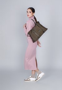 Olijfgroene nylon totebag met zilverkleurige accenten, opvouwbaar ontwerp en verstelbare straps, gedragen door een model in een roze outfit en sneakers.