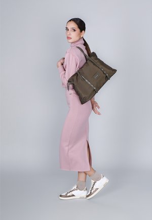 Olijfgroene nylon totebag met zilverkleurige accenten, opvouwbaar ontwerp en verstelbare straps, gedragen door een model in een roze outfit en sneakers.