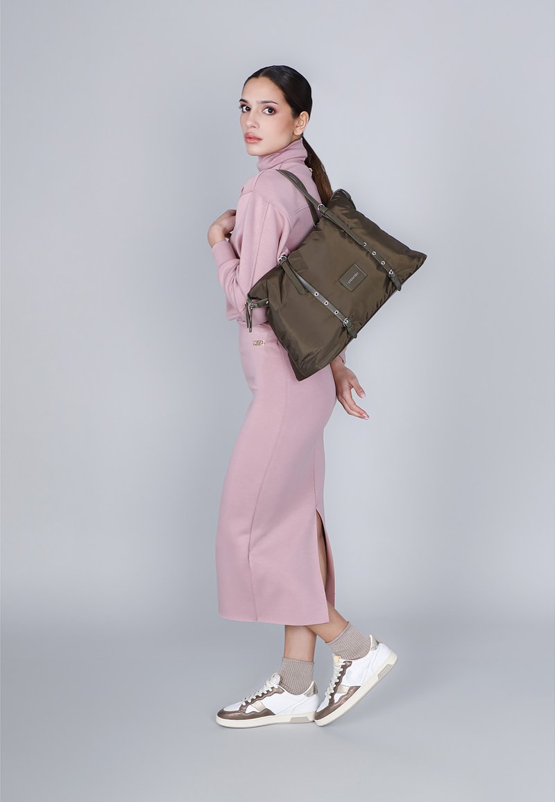 Olijfgroene nylon totebag met zilverkleurige accenten, opvouwbaar ontwerp en verstelbare straps, gedragen door een model in een roze outfit en sneakers.