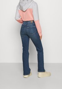 Kvinna iklädd blå denimjeans, ljusrosa och grå huvtröja, samt beige plattformssneakers, som står framför en ljus, enfärgad bakgrund.