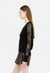 Pullover nero con corpo lavorato a maglia e maniche lunghe in pizzo, abbinato a una gonna di pelle plissettata color marrone. Presenta una sottile decorazione laterale.