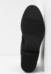 Bottine en daim noir avec bout rond, fermeture éclair latérale et semelle en caoutchouc texturée avec un logo. Dessus lisse avec des détails minimaux.