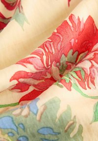 Blumestoff mit beigem Hintergrund, der große rote und rosa Blumen mit grünen Blättern und blauen Akzenten zeigt, aus einem weichen, strukturierten Material.