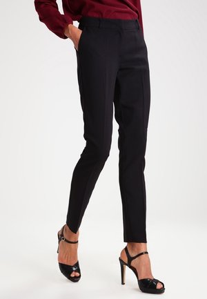 Mujer con pantalones negros ajustados, blusa de manga larga color burdeos y sandalias negras de tacón alto, de pie con una mano en el bolsillo.