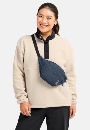 Glimlachende vrouw die een beige fleece trui en zwarte broek draagt, met een marineblauwe schoudertas met twee ritsvakken op de borst.