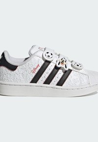 Zapatillas blancas con parte superior texturizada que presenta un patrón de personajes animados. Diseño de tres rayas negras, suela de goma y detalles únicos en el cierre.