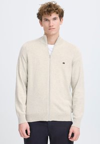 Suéter de cremallera color beige claro con un cuello alto, puños y dobladillo acanalados, que presenta un pequeño logo negro en el pecho. Textura suave y de punto.