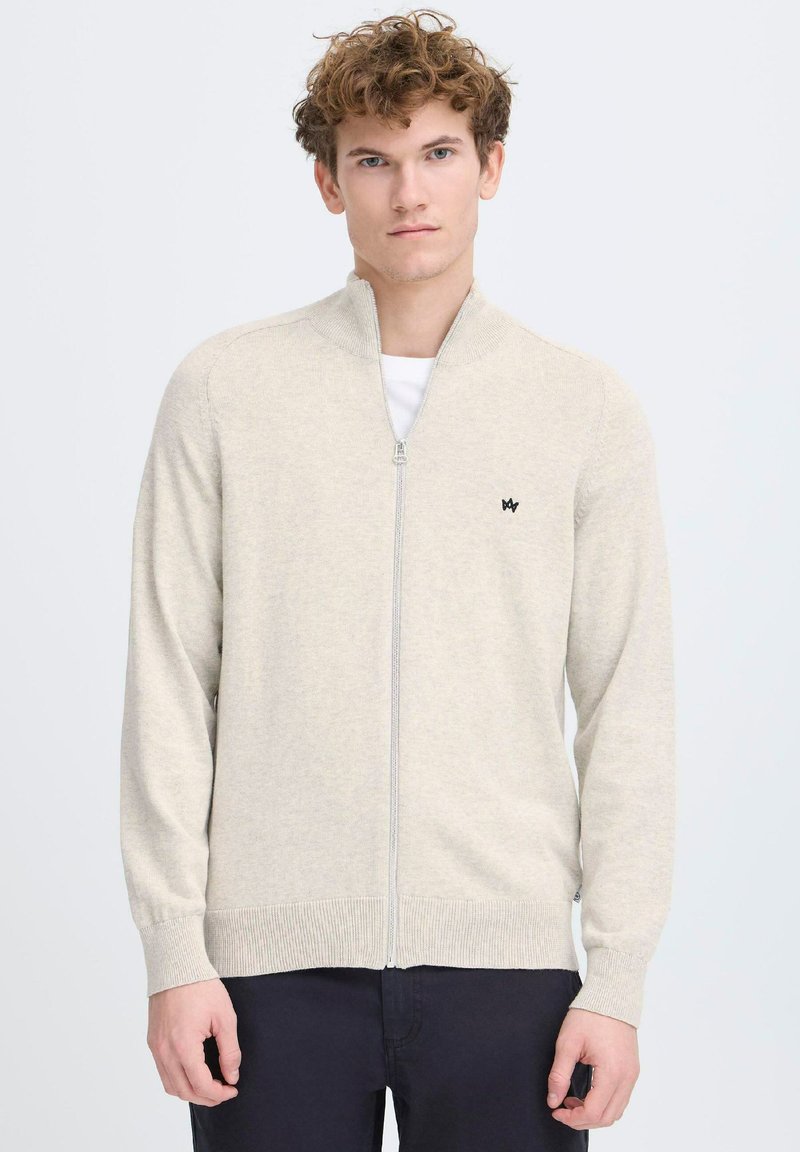 Suéter de cremallera color beige claro con un cuello alto, puños y dobladillo acanalados, que presenta un pequeño logo negro en el pecho. Textura suave y de punto.