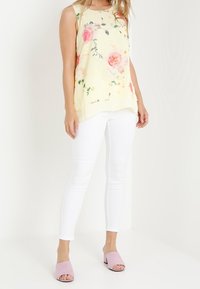 Žlutý top bez rukávů s květinovým vzorem, doplněný bílými úzkými kalhotami a růžovými sandály na podpatku. Hladká látka, volný střih.