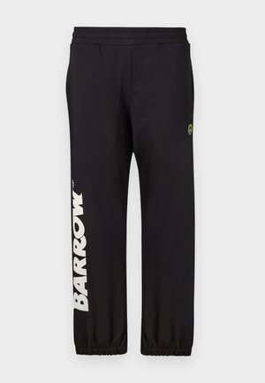 Ung mand står med hænderne i lommerne iført sort t-shirt og sweatpants med gule smilende ansigtslogoer og hvide sneakers på ensfarvet baggrund.