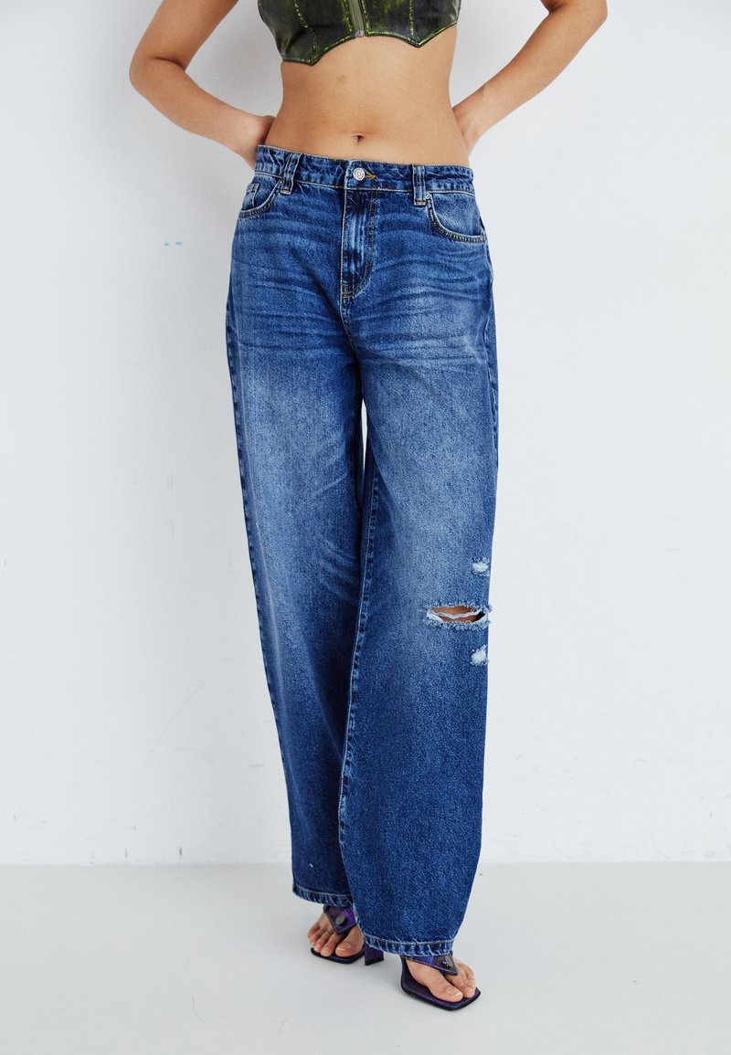 Højtaljede, løse blå denimjeans med slidte huller. Har et falmet look, fem-lomme design og sølvfarvet knaplukning.