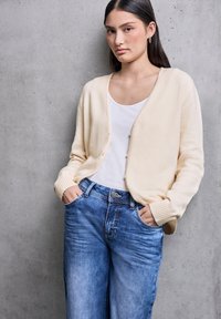 Cardigan lavorato a maglia color crema con collo rotondo, apertura con bottoni e polsini a costine, indossato sopra una camicia bianca e jeans a gamba larga blu.